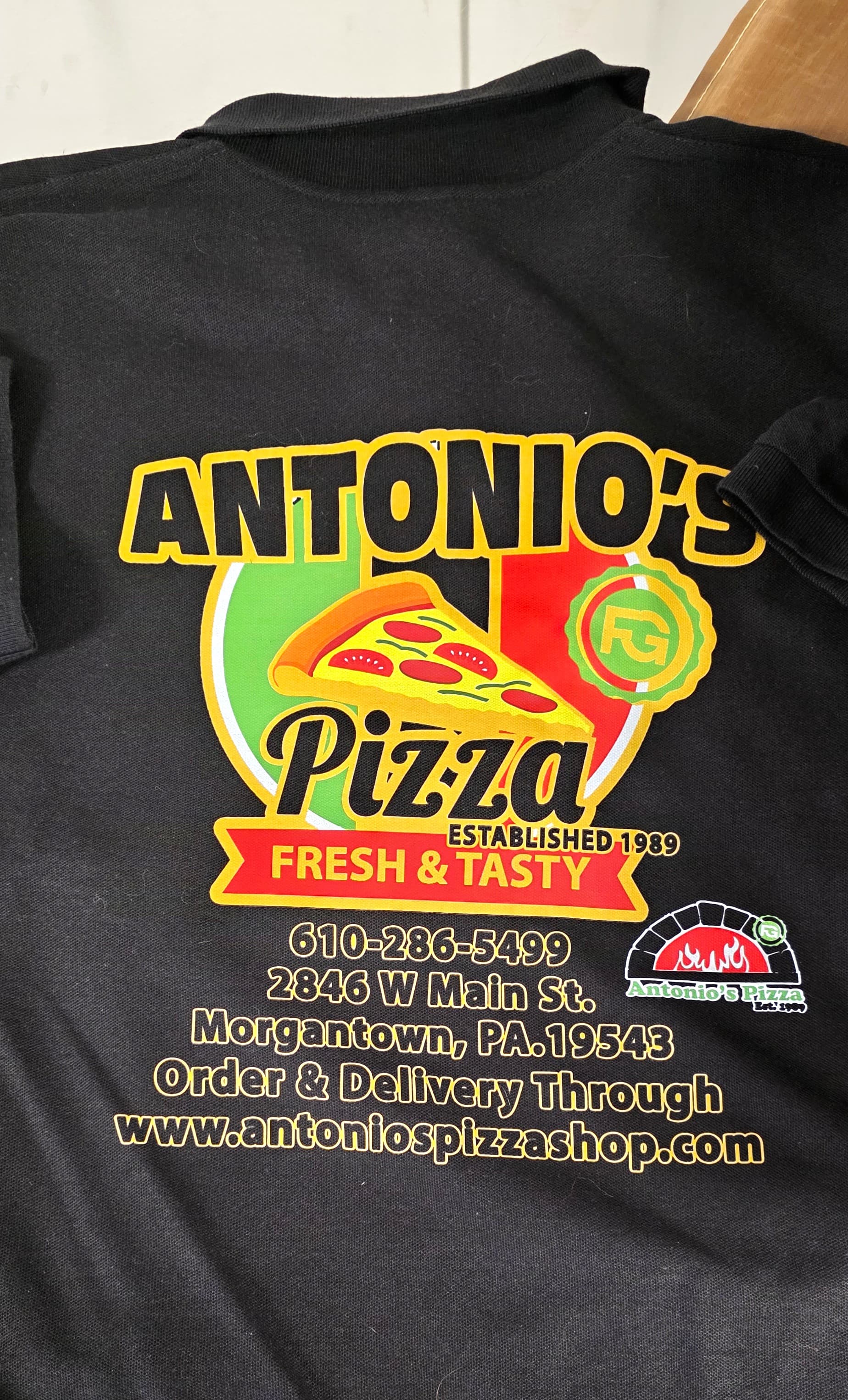 Full-color back print on a black Antonio's Pizza polo — Fresh & Tasty, Est. 1989, with phone, address, and website — by Print Habit