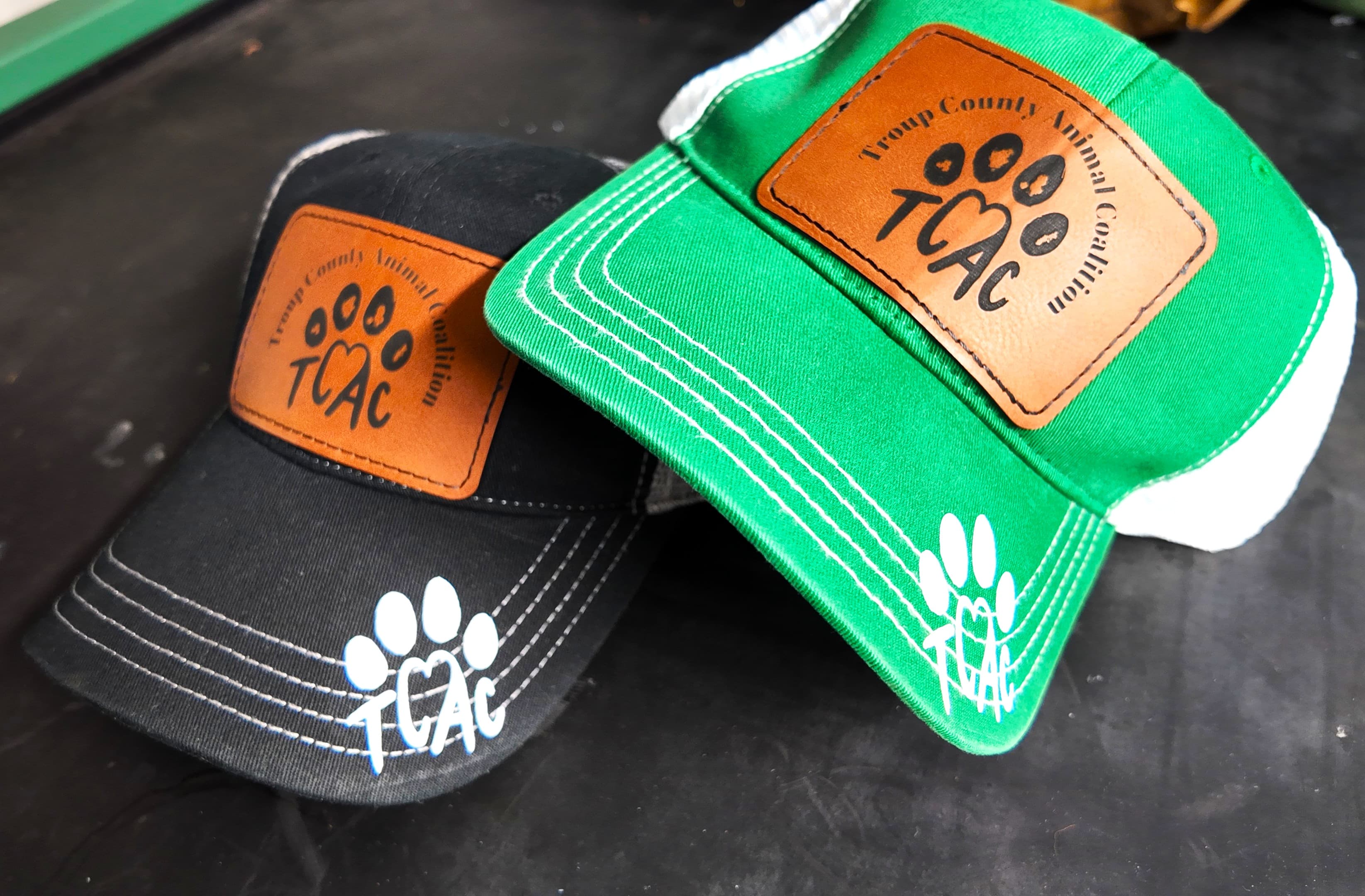 Custom black and green trucker hats with laser-engraved leather TCAC patches and a printed paw print on the bill — Troup County Animal Coalition, by Print Habit